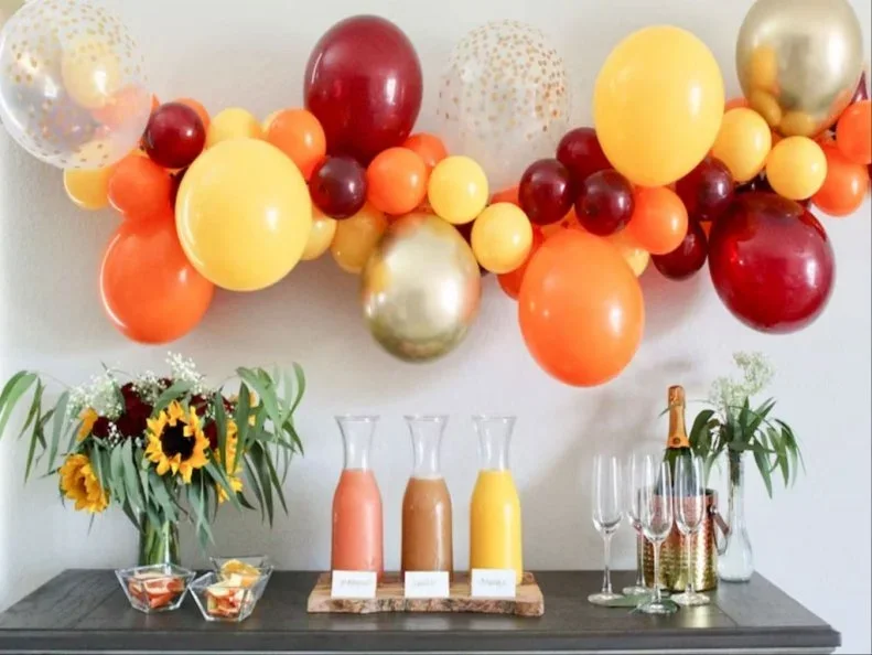 Orange, yellow, red and warm metallic balloons form a garland above a dark drinks surface with flowers and glass bottles