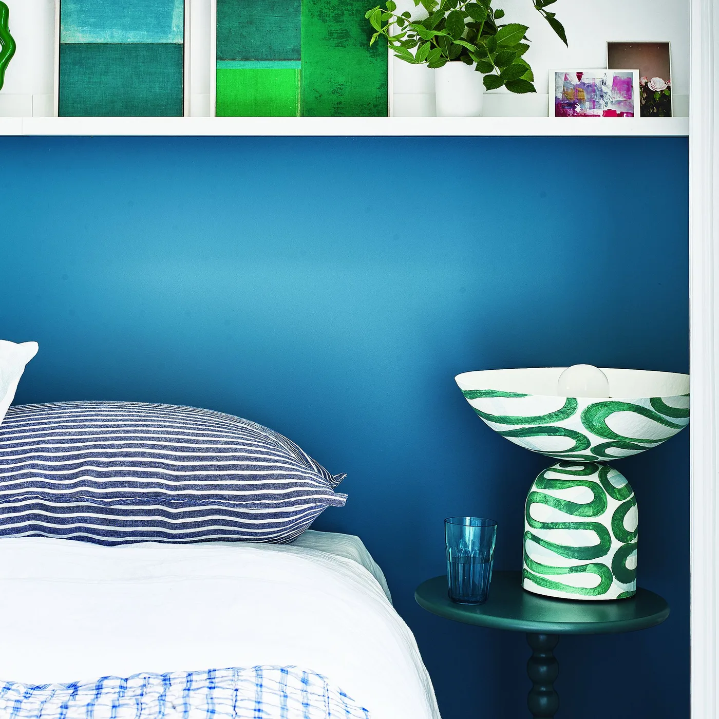 Blue bedroom wall with a narrow white display shelf, green abstract pictures, striped pillow and a green-and-white bowl on a small table.