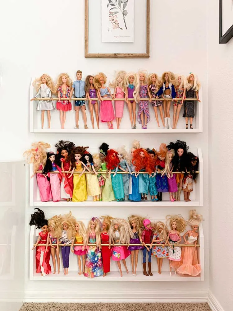 Three white wall racks with gold-colored retaining bars holding rows of fashion dolls.