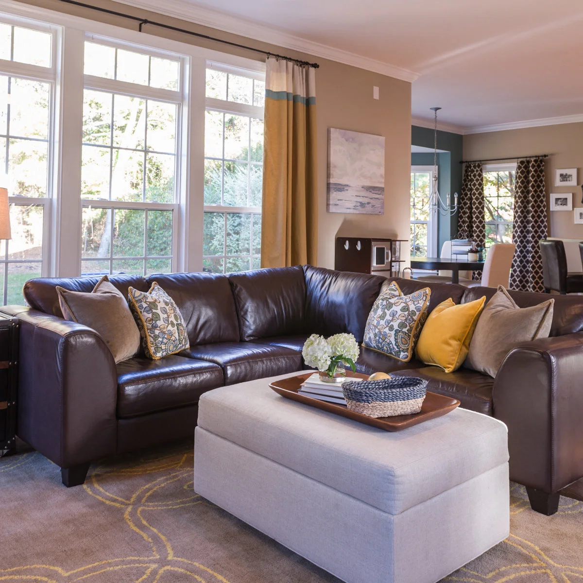 Dark brown leather sectional with yellow curtains, pale patterned cushions and a light gray ottoman.