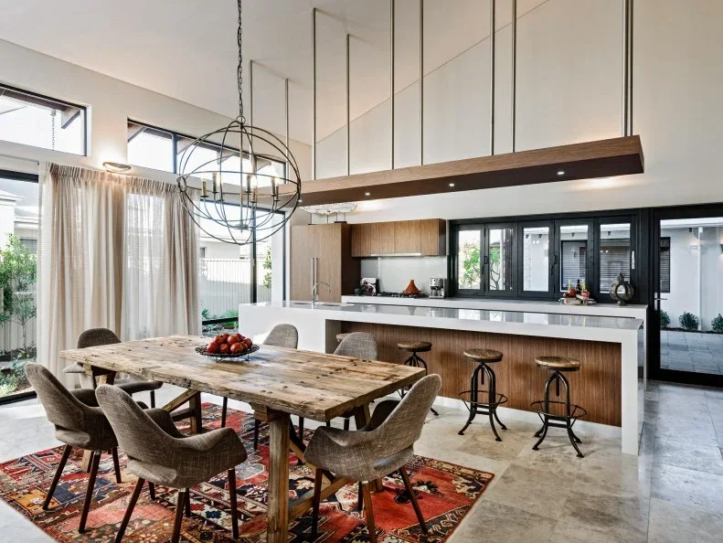 A rough wood dining table and upholstered chairs sit on a red patterned rug beside a white kitchen island.