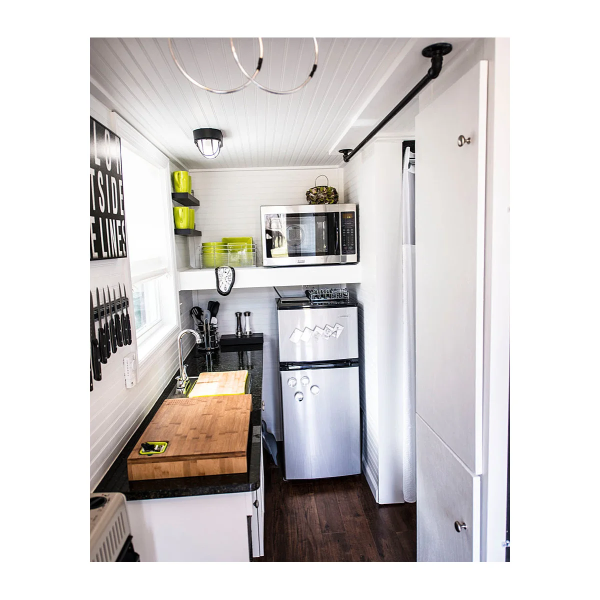 Tiny white kitchen with cutting boards beside a small sink and compact refrigerator at the end