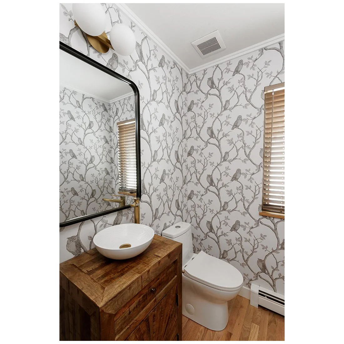 Gray branch-and-bird wallpaper surrounds a wood vanity with a white vessel bowl, large dark-edged mirror, warm-metal tap and pale wood floor.