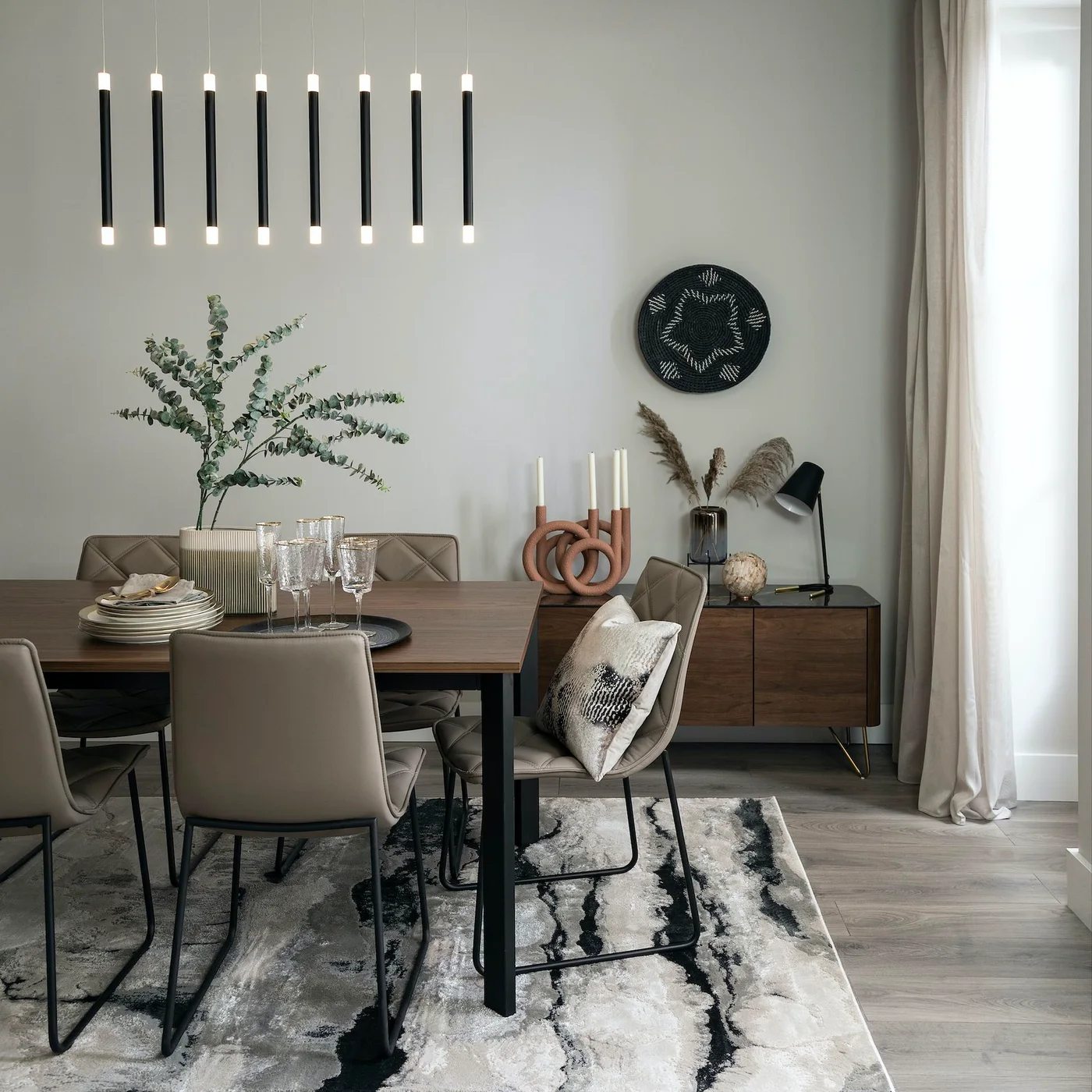 A dark wood table and slim-legged upholstered chairs stand near a low wood sideboard, pale curtains and a black-and-white patterned rug.