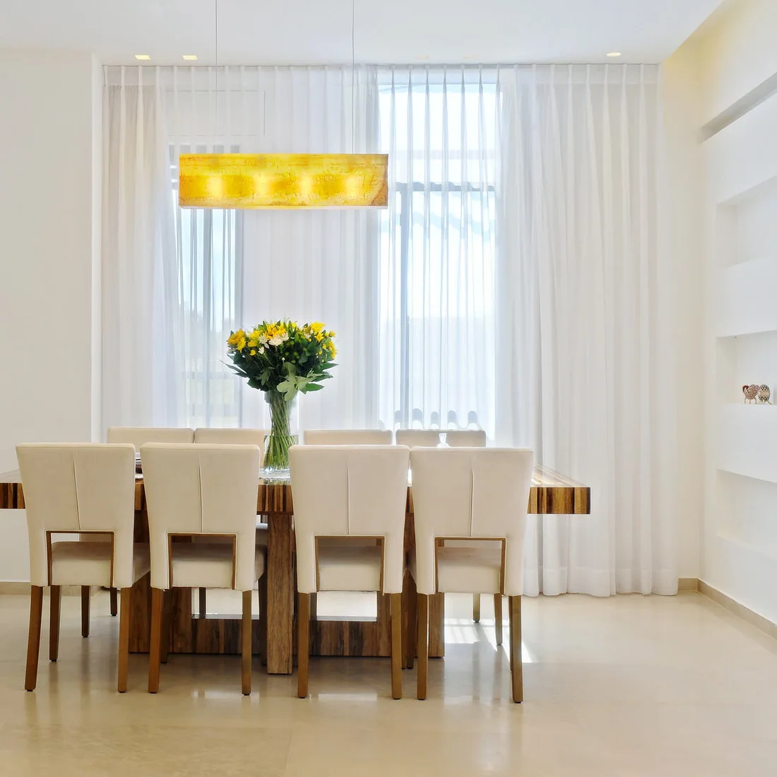 A wood dining table and tall pale upholstered chairs stand beneath a yellow rectangular hanging light beside white curtains and yellow flowers