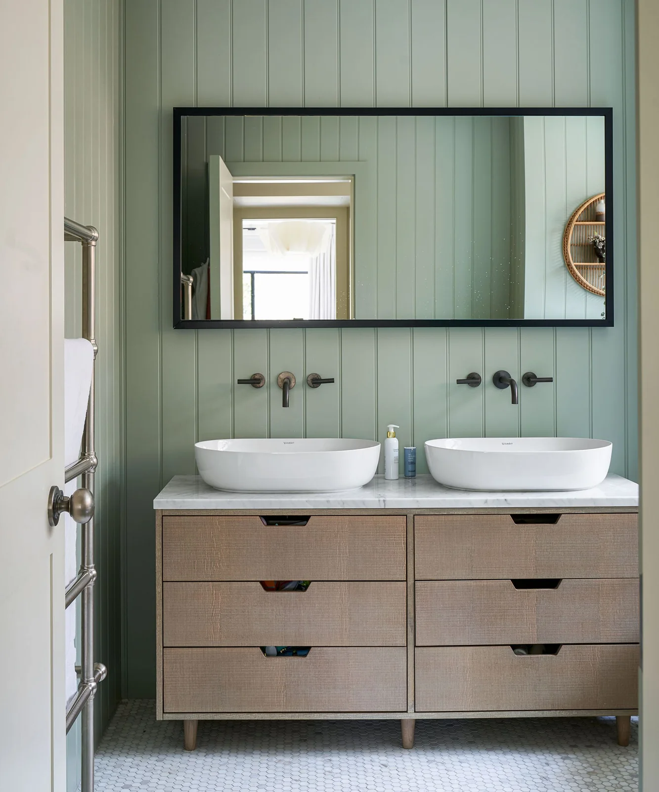 Two white oval basins sit on a wood drawer vanity beneath a broad black-framed mirror against a pale green vertically paneled wall