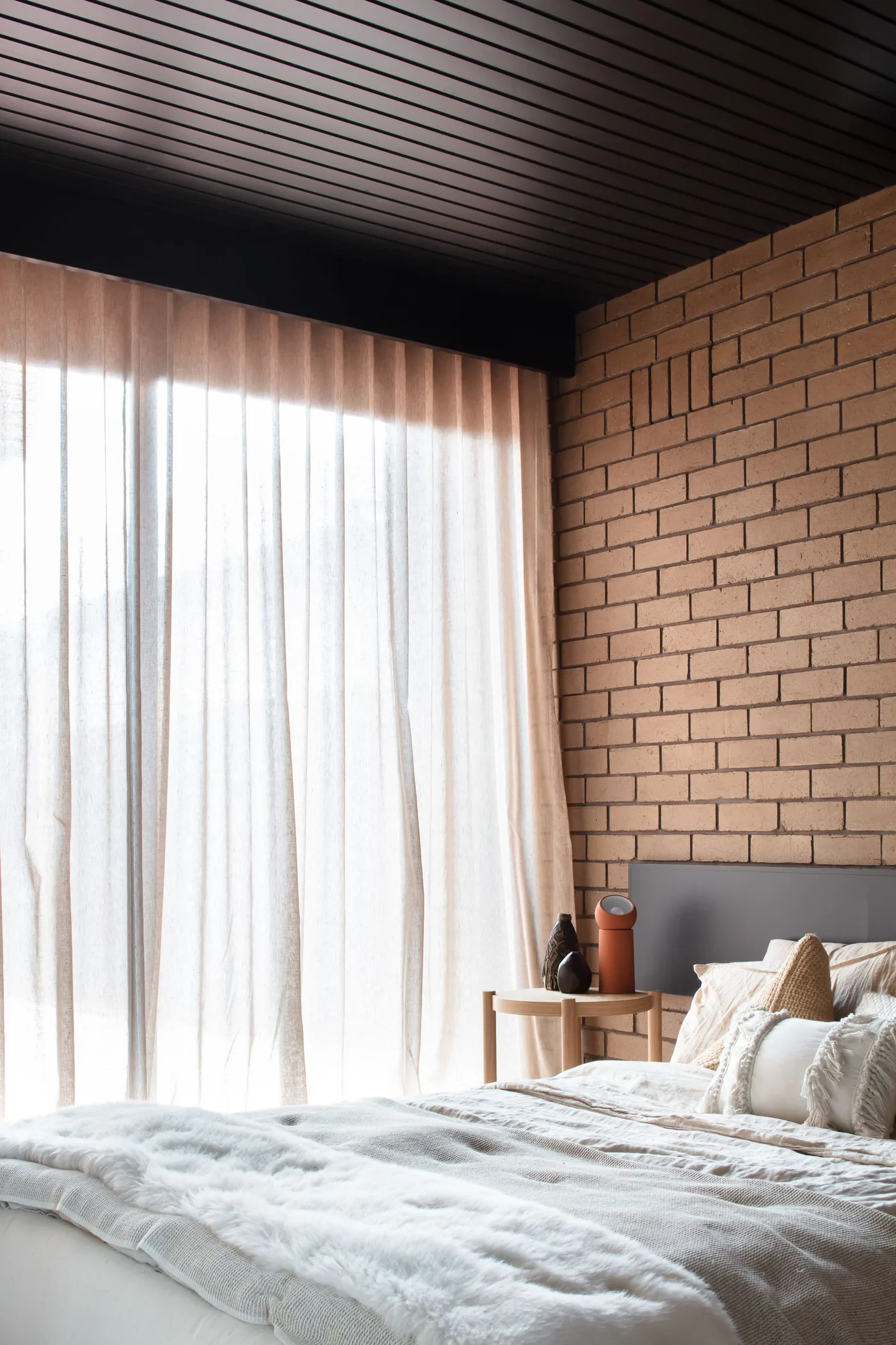 Bedroom with exposed-looking brick walls, a dark slatted ceiling and pale bedding beside sheer curtains