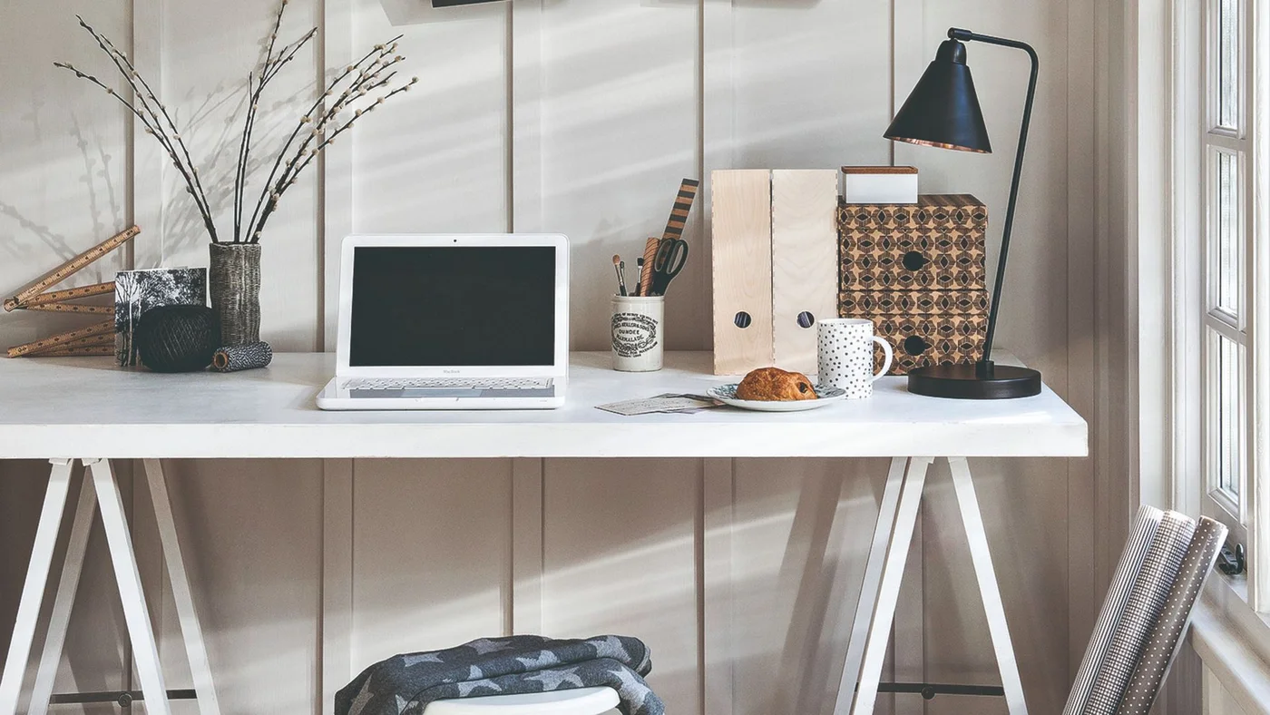 A white trestle desk stands against pale vertical wall boards beside a window, with a black task lamp, a small laptop and upright document files.