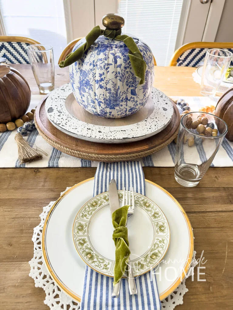 Cutlery tied with a green ribbon rests on a striped napkin above layered pale plates beside a blue-and-white pumpkin