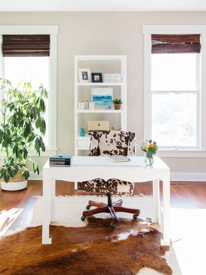 White desk with a brown-and-white patterned rolling chair, layered rugs and bright windows.