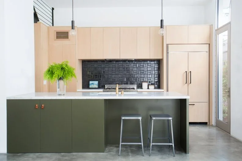An olive green flat-front island stands before pale wood cabinets and a dark patterned backsplash, with two simple metal stools.