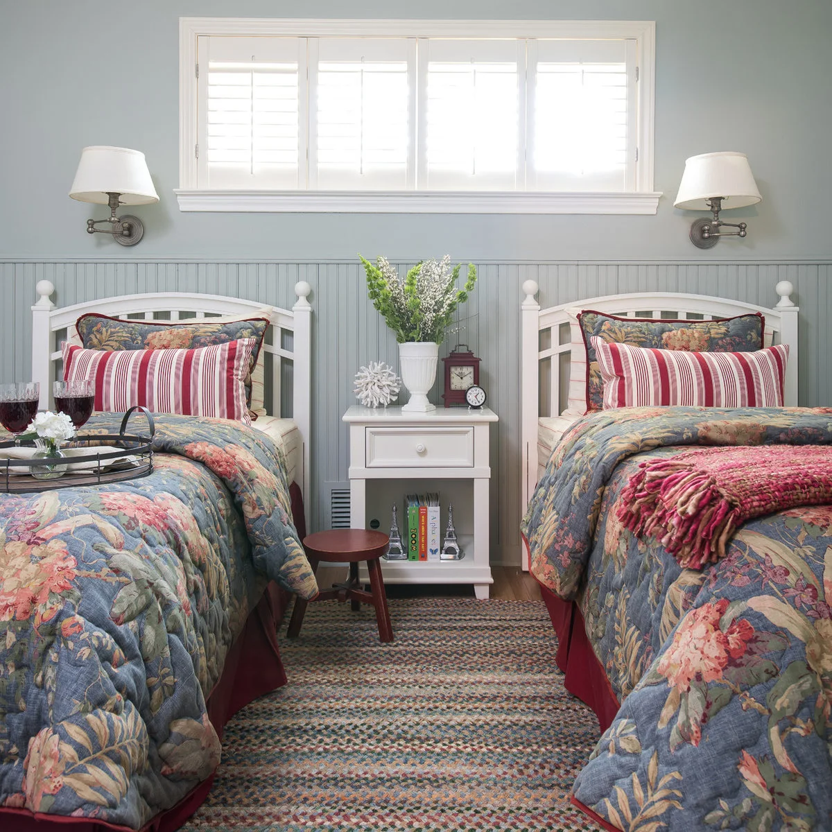 Two beds with floral covers, striped cushions, a small white bedside table and wall lamps above