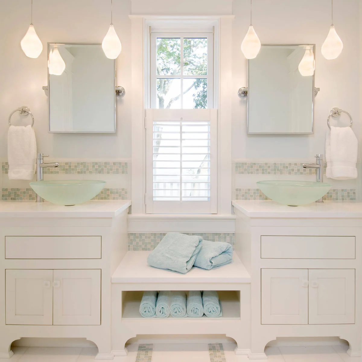 Two pale aqua translucent vessel bowls sit on white vanities separated by a lower towel shelf beneath a tall window.
