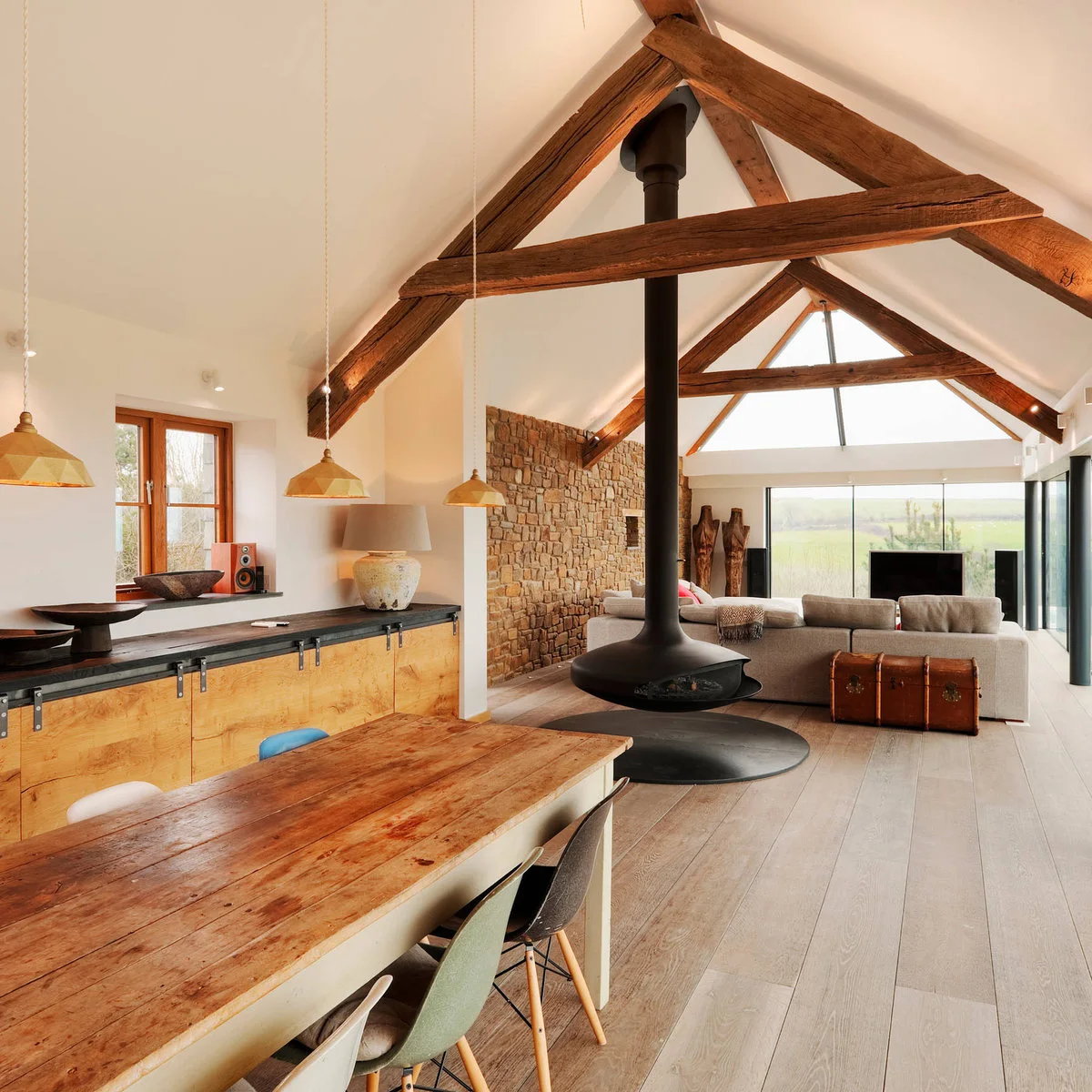 Long beamed room with a wooden dining table, suspended dark fireplace and gray sofas facing the glazed end.