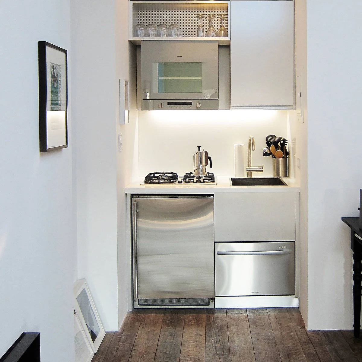 Small black-framed picture on the side wall beside a compact white kitchenette with stainless appliances.
