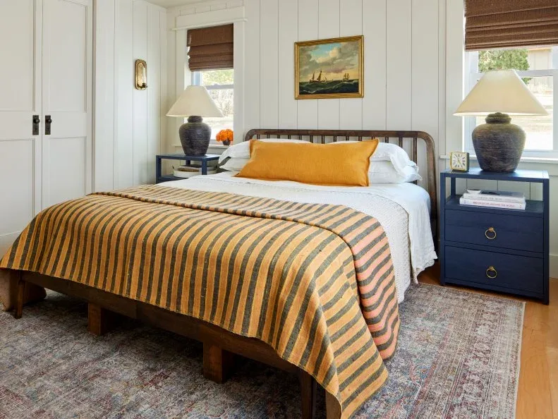 White bedroom boards surround a timber bed with an ochre striped cover, dark blue drawers and broad shaded lamps.