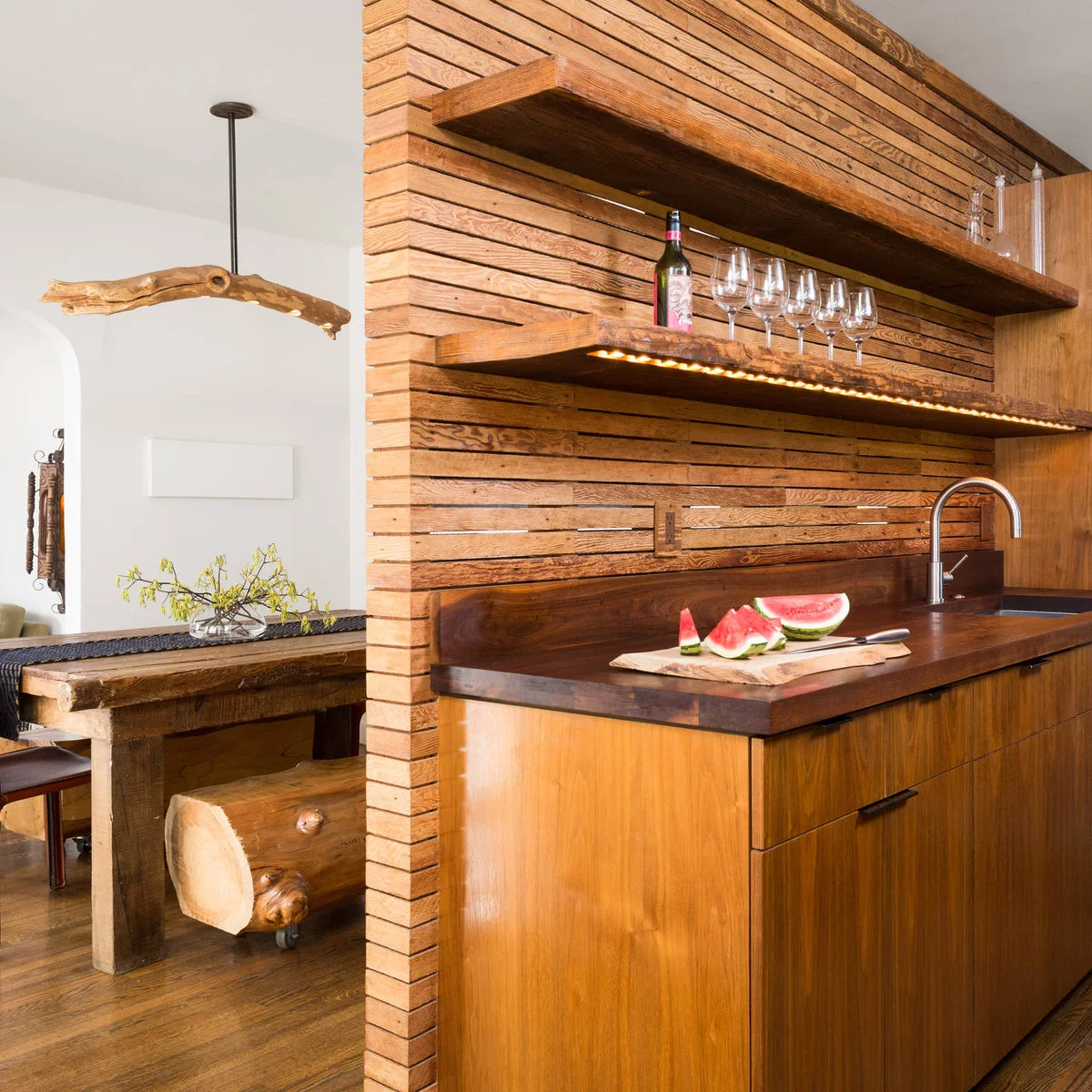 A horizontal wood slat divider supports shelves above a sink counter, with a dining table visible beyond.