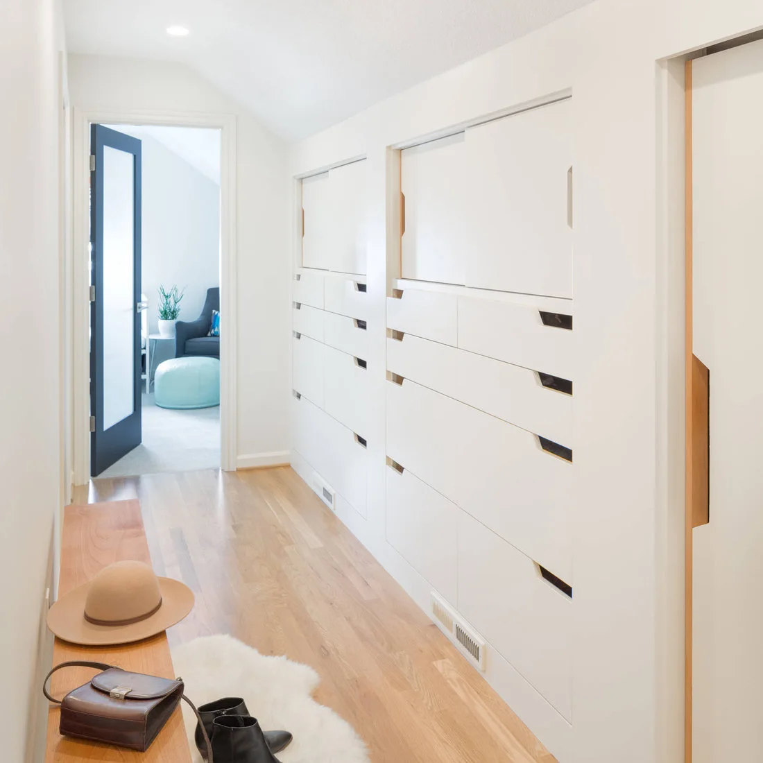 White sliding storage panels and rows of drawers line a pale hallway opposite a slim wood bench beneath a blue doorway.