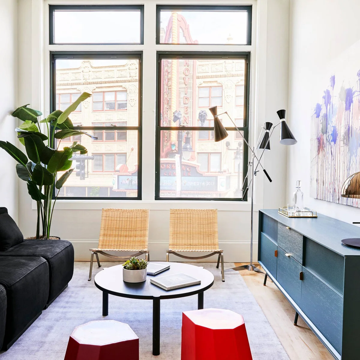 Small white-walled room with dark seating, woven chairs, a blue sideboard and red stools