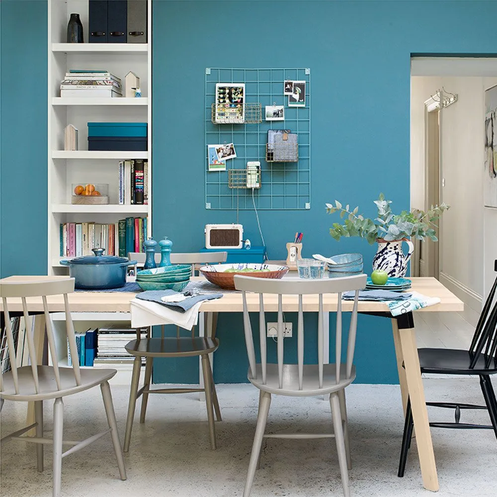 Narrow white fitted bookcase beside a teal dining-room wall, with books and boxes, a wooden table, mixed chairs, and a wire organizer.