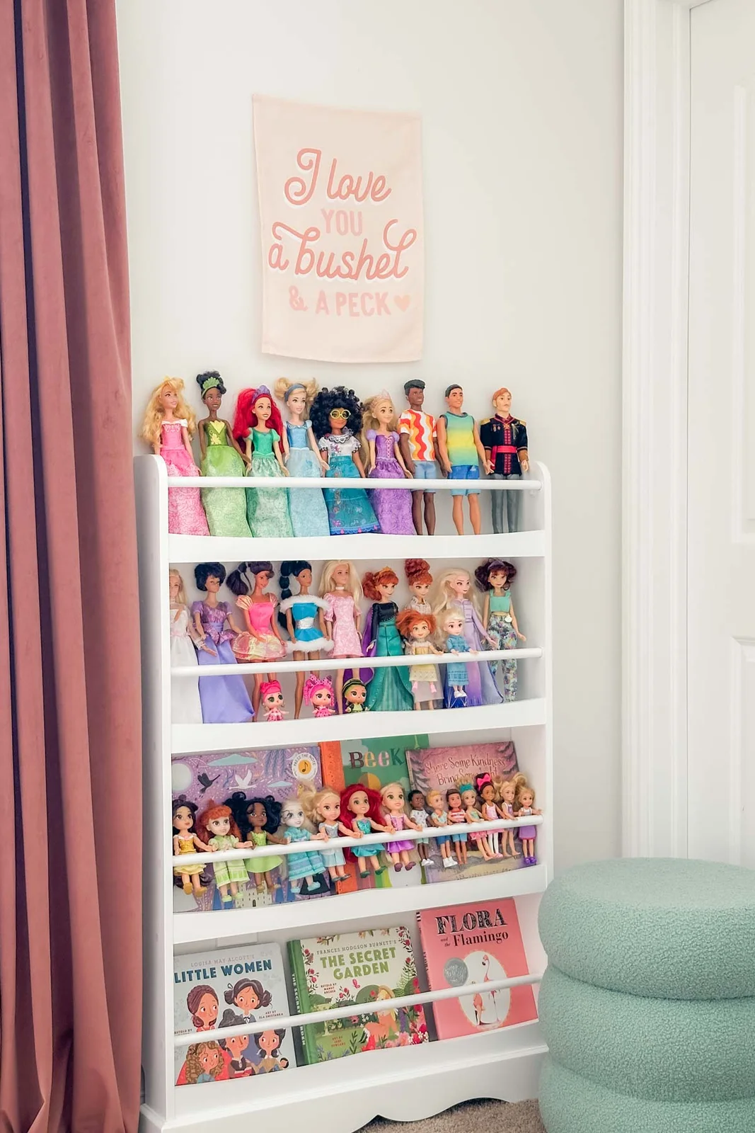 White forward-facing book rack holding fashion dolls, smaller dolls and picture books beside a mint ottoman.