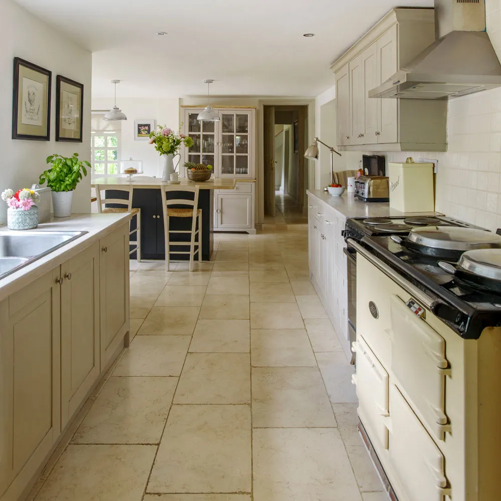 Cream paneled cabinets line a long kitchen beside pale counters, a dark range and a tiled floor