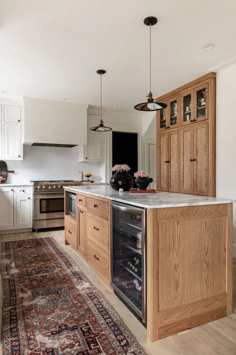 A wood island with drawers and a wine refrigerator stands beside a tall wood cabinet, with white cupboards around the range.