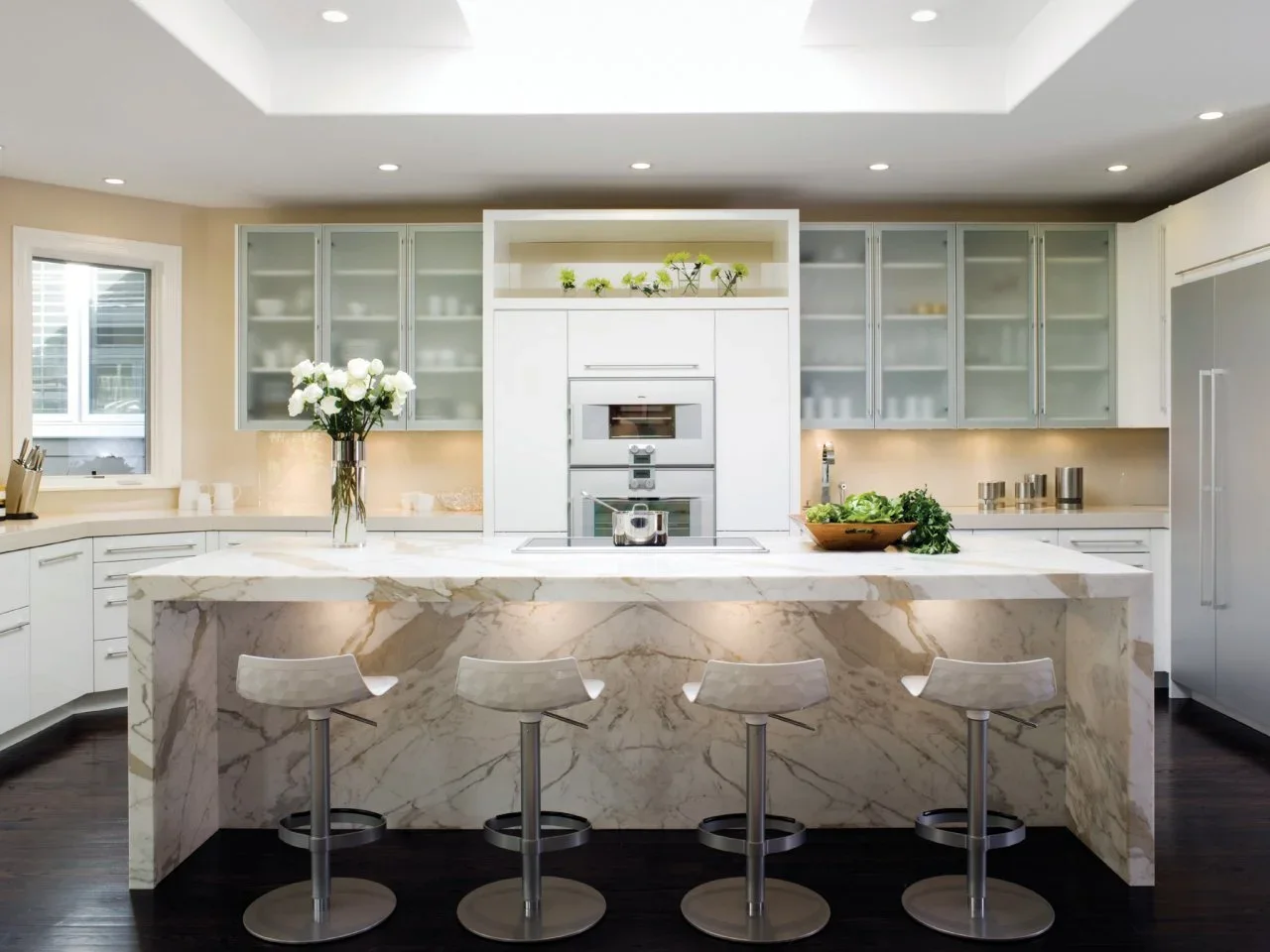 White cabinets with frosted glazed upper doors surround an oven tower beside a large veined island.