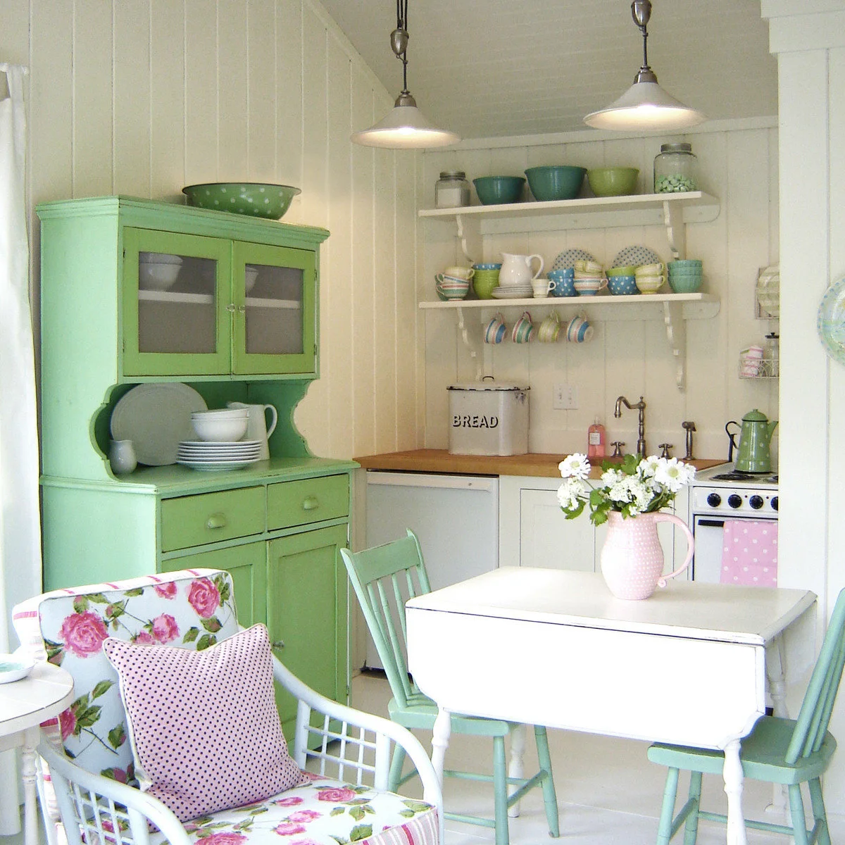A bright green hutch and mint chairs sit in a cream-paneled kitchen with a white table, pink accessories and floral upholstery.