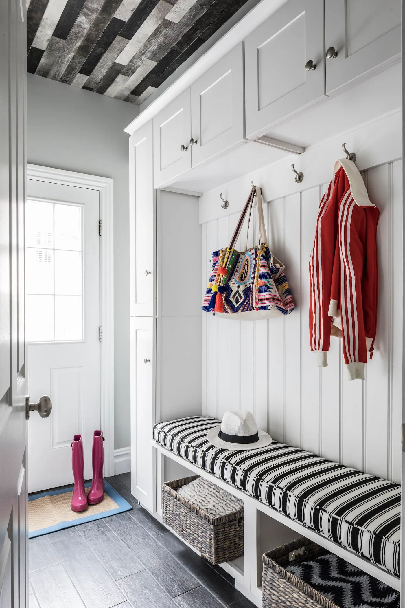 White paneled mudroom bench with black-and-white striped upholstery, overhead cupboards and baskets below.