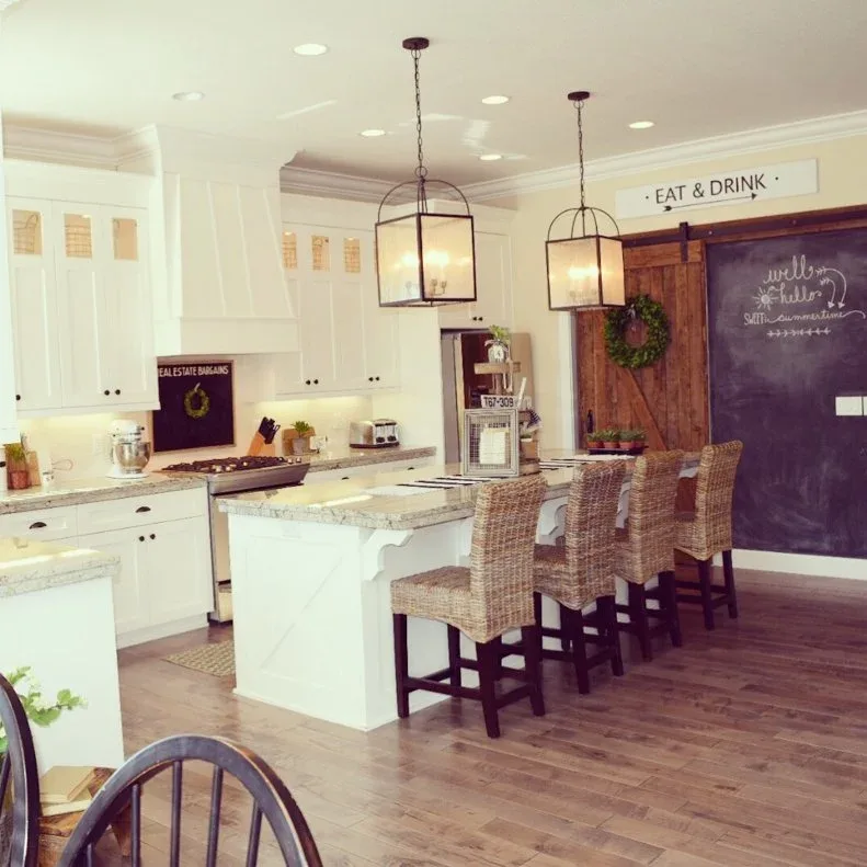 White country kitchen with woven bar stools, lantern pendants and a dark chalkboard wall beside a sliding wood door