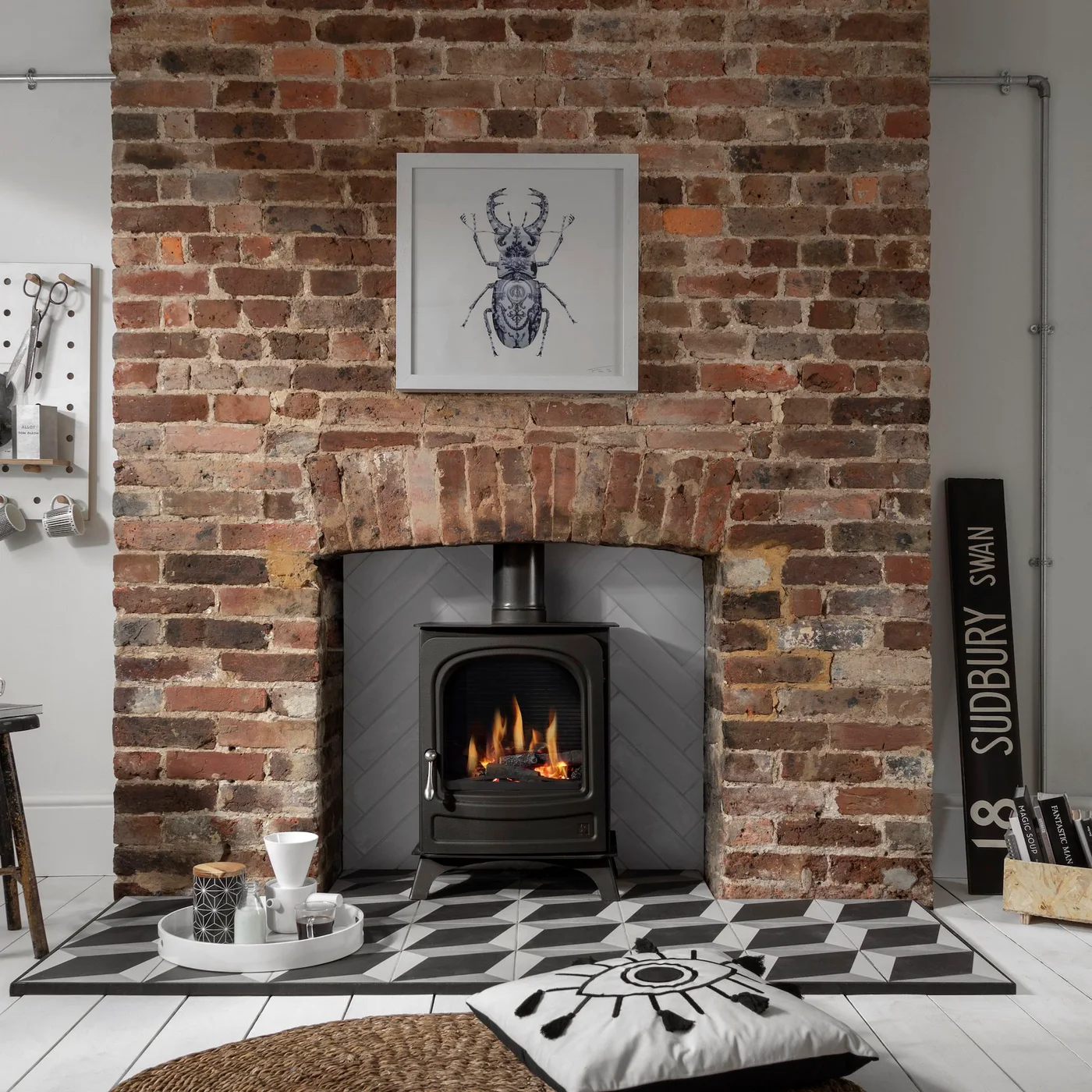 Gray herringbone tile lines an exposed brick fireplace behind a black stove above a black-and-white geometric tiled hearth.