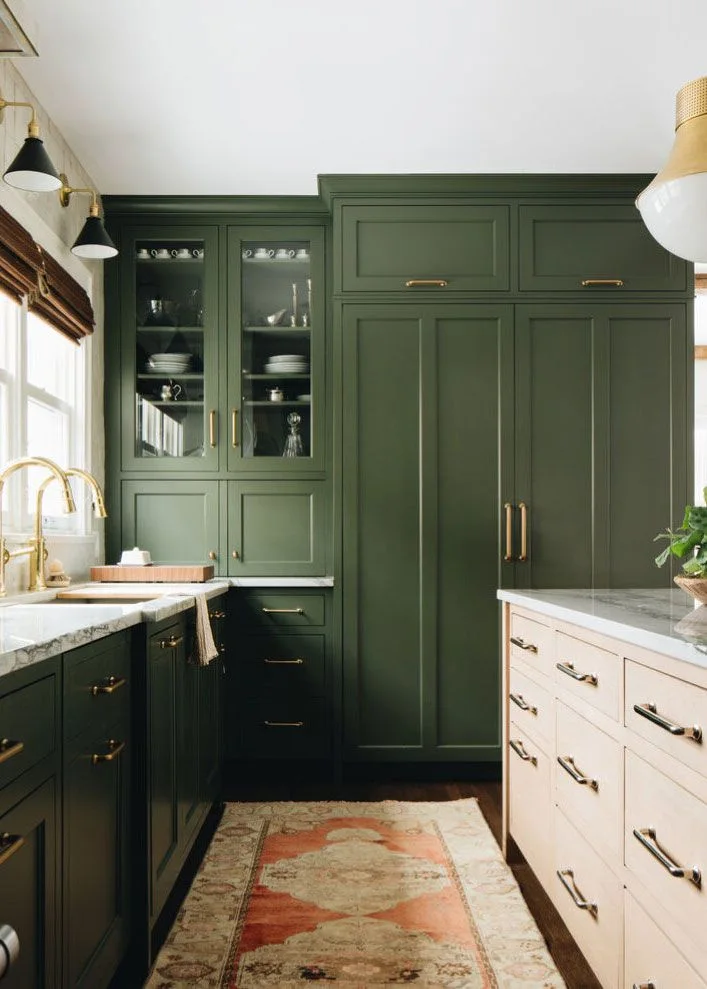 Pale cream kitchen island drawers opposite deep green cabinets, warm-metal hardware, glass-fronted storage and a patterned runner.