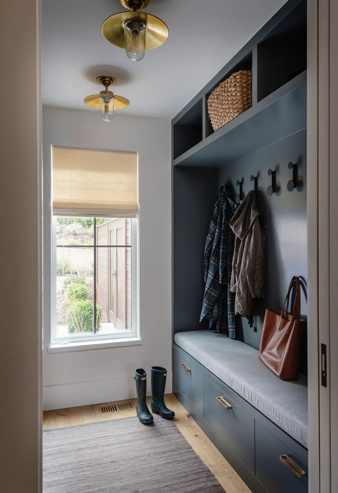 A muted blue built-in bench has a gray seat, dark coat hooks, drawers and upper cubbies beside a window with a pale blind