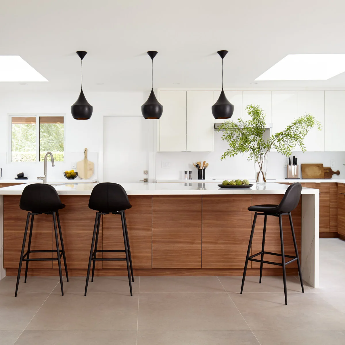 Black curved stools and three black pendants against a pale kitchen with wood island fronts and white upper cabinets.