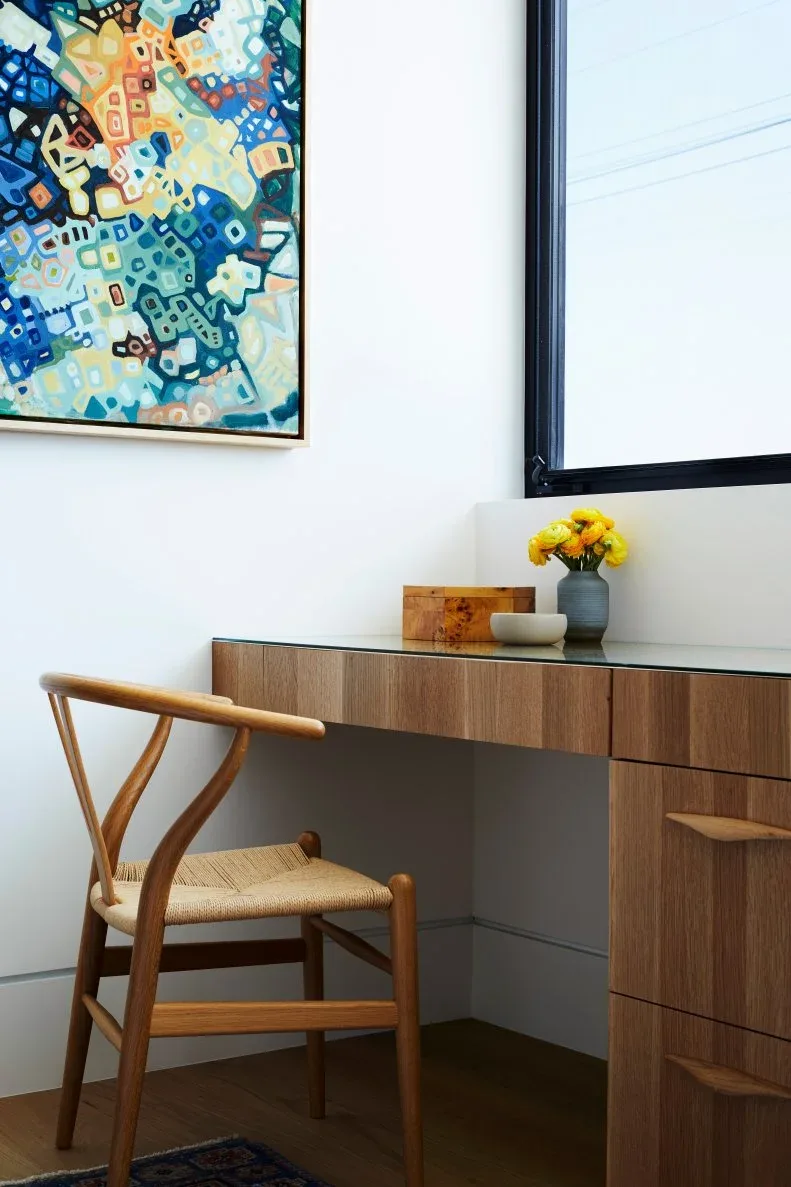 A wood drawer desk with a dark top sits beside a black-framed window, with a curved wood chair, colorful abstract artwork and a small vase of yellow flowers.