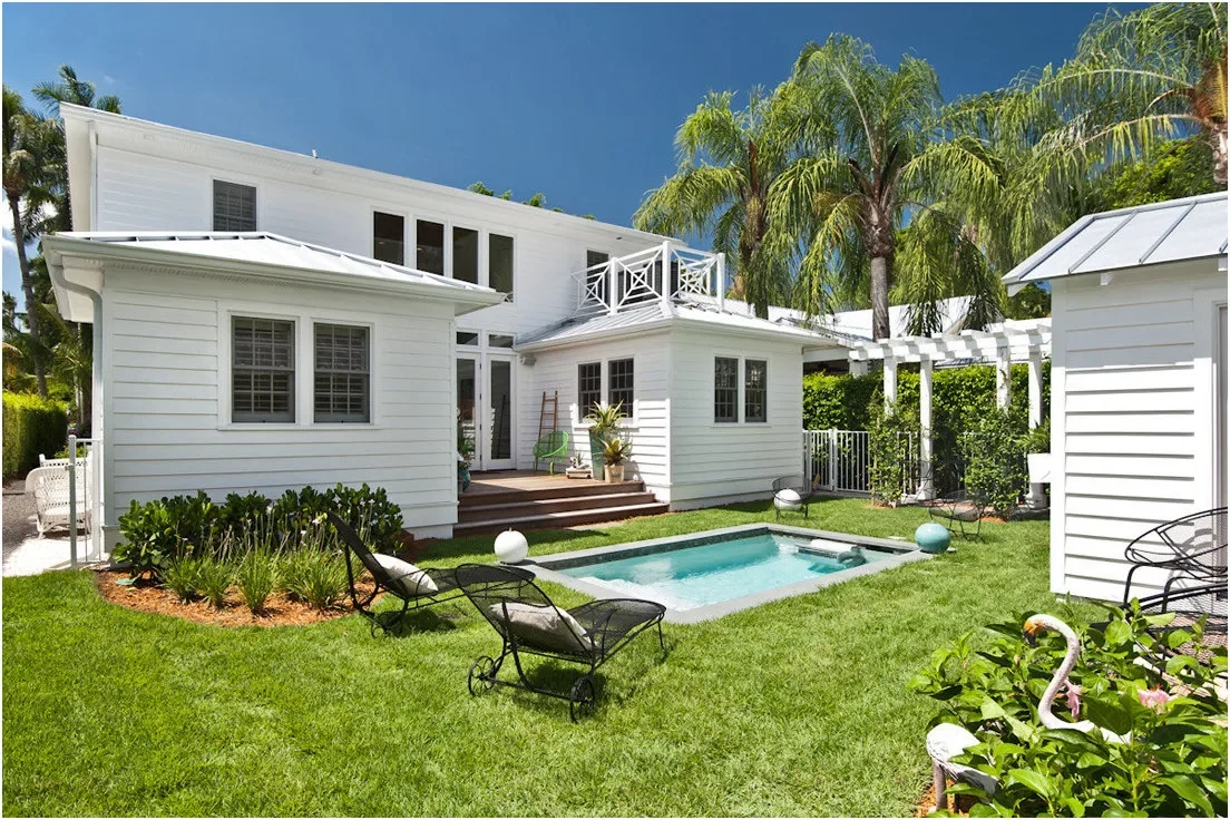 A small rectangular pool sits in a lawn courtyard beside a white house, pale paving and dark outdoor chairs.