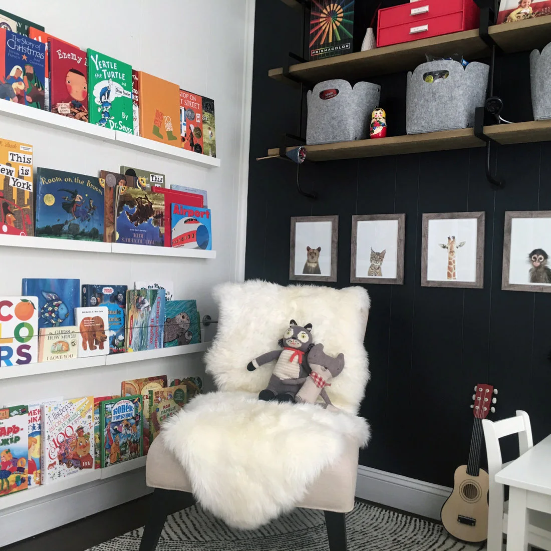 A reading corner has white face-out book ledges, a white fluffy chair, black wall paneling, wood shelves with gray bins and small animal pictures.