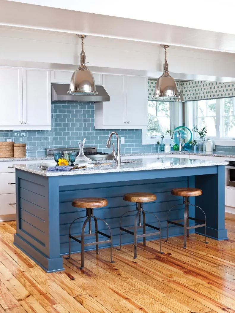 A blue paneled island with wood-topped stools stands beneath metal pendant lights beside white cupboards and blue rectangular backsplash tiles