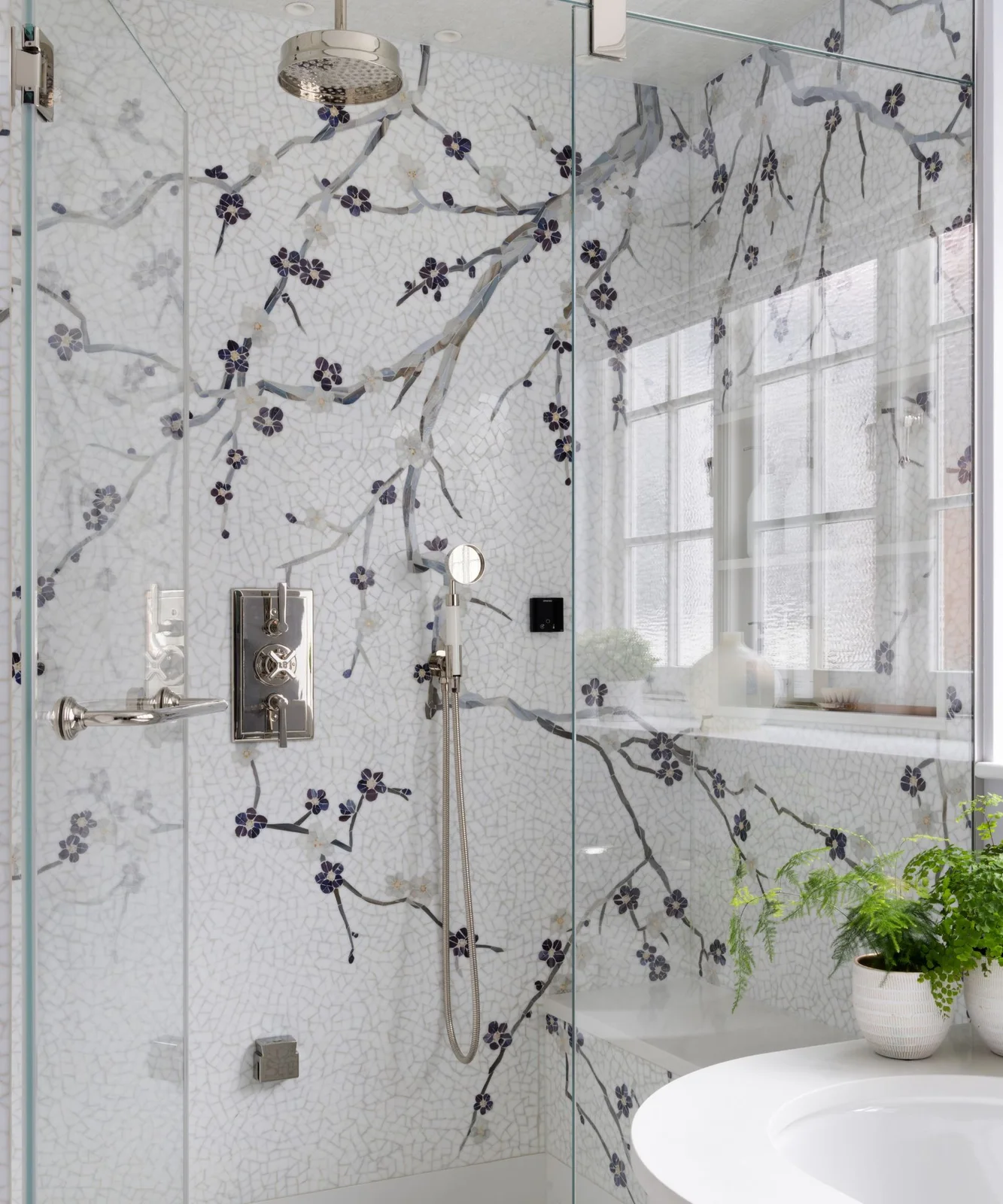 A flowering-branch mosaic passes behind a shower head, controls and hand shower beside a glass screen.