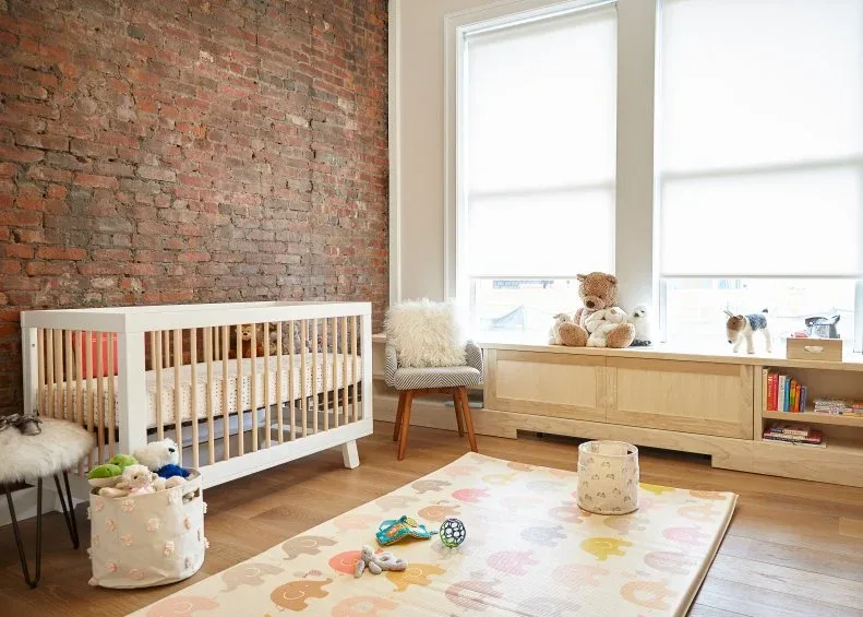Nursery with exposed brick wall, pale wood crib and low storage, large windows, colorful rug and small chair