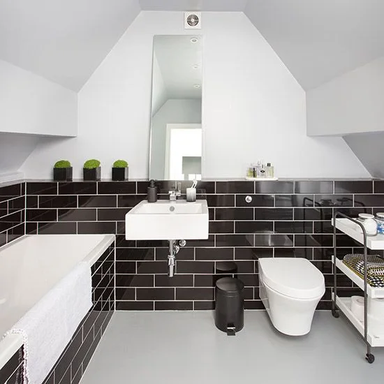 An attic bathroom has glossy dark lower wall tiles, white sloped ceilings, a white rectangular basin and a tall mirror beside a bath and toilet