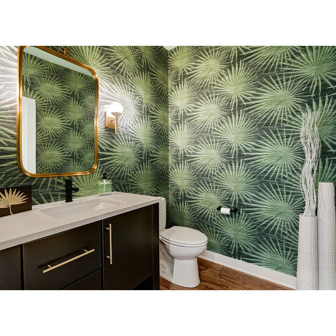 Pale green radiating leaf wallpaper surrounds a dark powder-room vanity and gold-colored mirror beside a white toilet and tall decorative branches