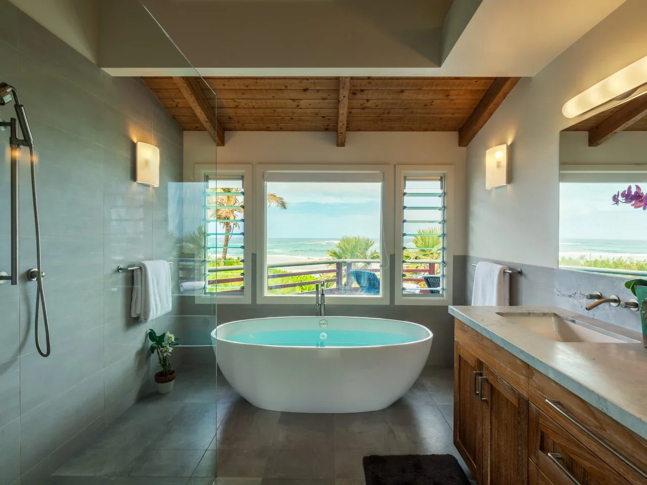 A timber ceiling and vanity warm a gray bathroom with a white tub, glass shower and wide window.