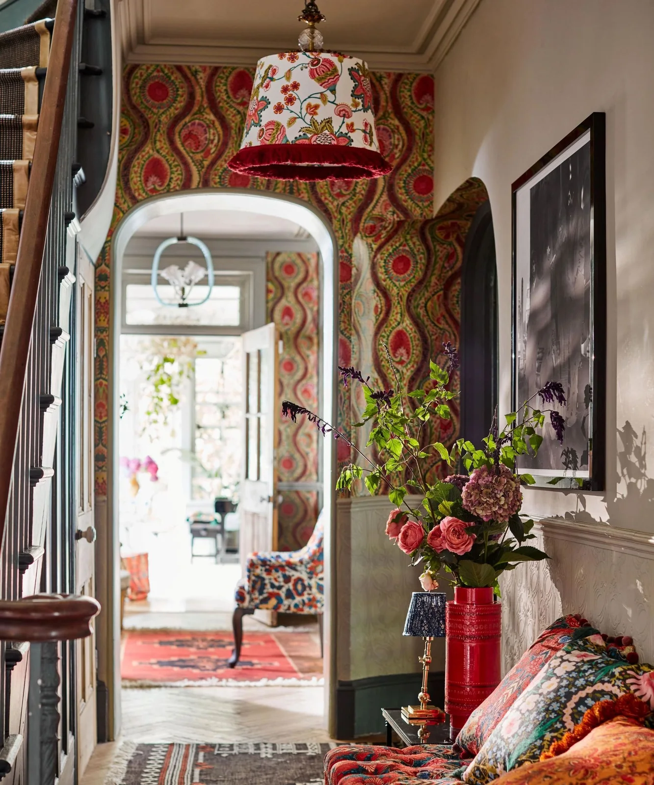 A patterned pendant hangs above floral cushions and a bench beside red-and-green patterned wallpaper, colorful rugs and a dark stair railing