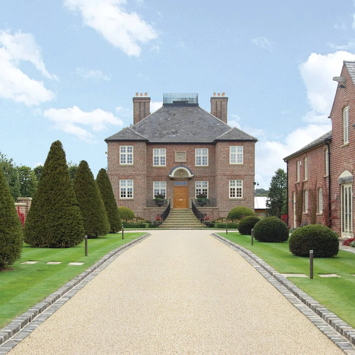 Straight pale driveway toward a brick house, edged with low blocks, lawn, clipped shrubs and slim path lights.