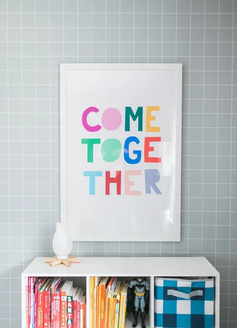Pale gray grid wallpaper stands behind a white-framed colorful print above a low white cubby unit holding books and a blue checked basket.