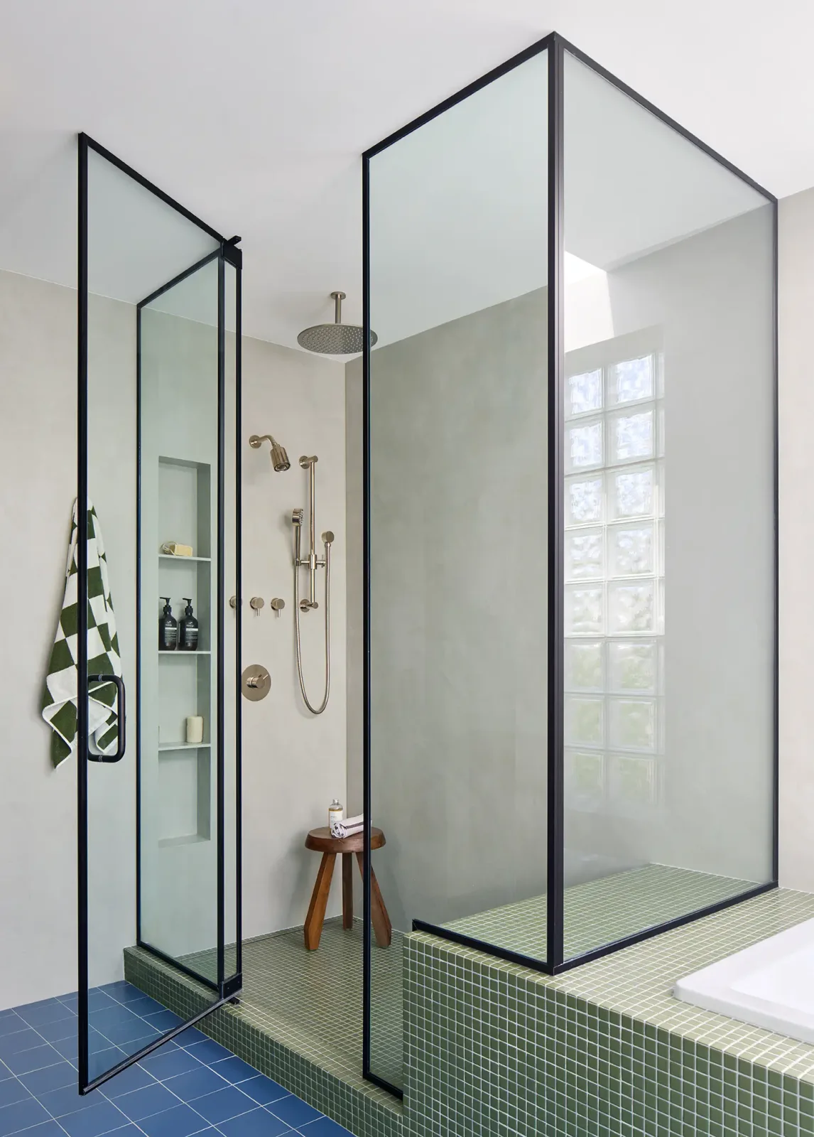 A black-framed glass shower stands on an olive mosaic platform beside blue floor tiles, white walls and a glass-block window.