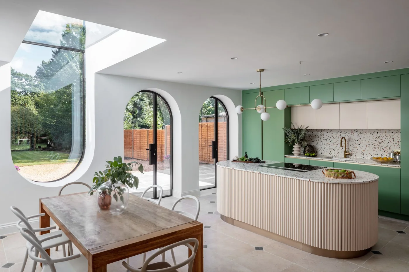 A wooden dining table stands beside a rounded fluted island, green cabinets and tall arched windows in a pale kitchen