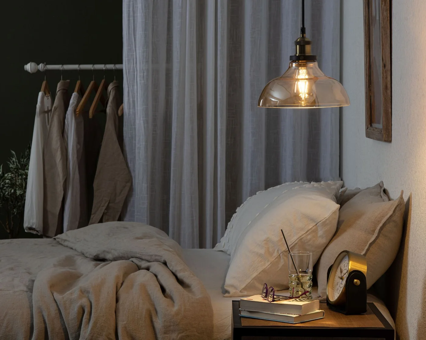 A glowing glass-shaded pendant hangs above pale bedding and a bedside surface with books beside open clothes storage and a white curtain